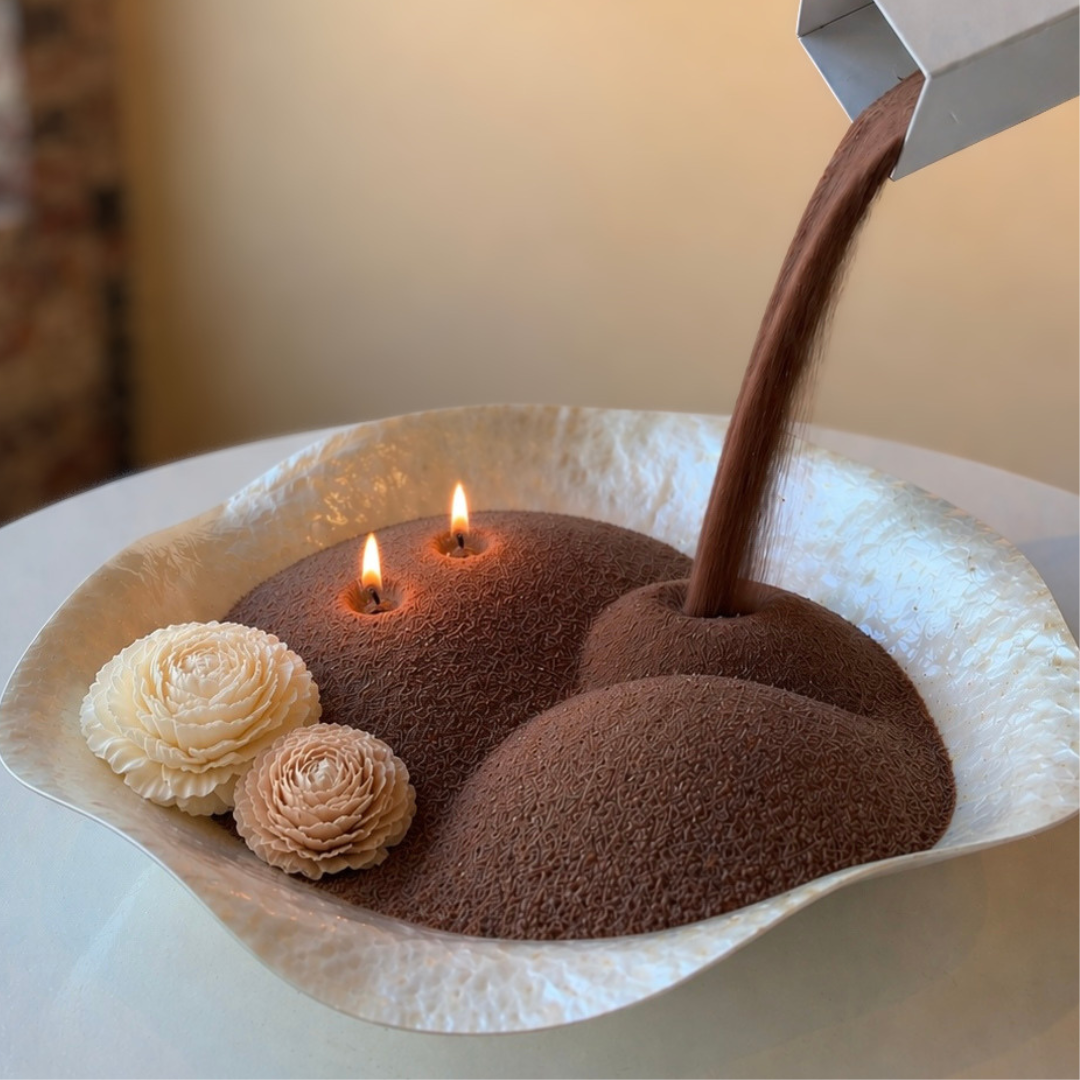 A visually satisfying lifestyle shot capturing the refill process of a pearled candle, as rich pecan wax pearls are poured into a sculptural bowl with softly glowing flames. Accented with delicate wax floral details, the scene blends artistry and function, highlighting the customizable, refillable nature of the candle while creating a warm, sensory experience.