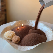 A visually satisfying lifestyle shot capturing the refill process of a pearled candle, as rich pecan wax pearls are poured into a sculptural bowl with softly glowing flames. Accented with delicate wax floral details, the scene blends artistry and function, highlighting the customizable, refillable nature of the candle while creating a warm, sensory experience.