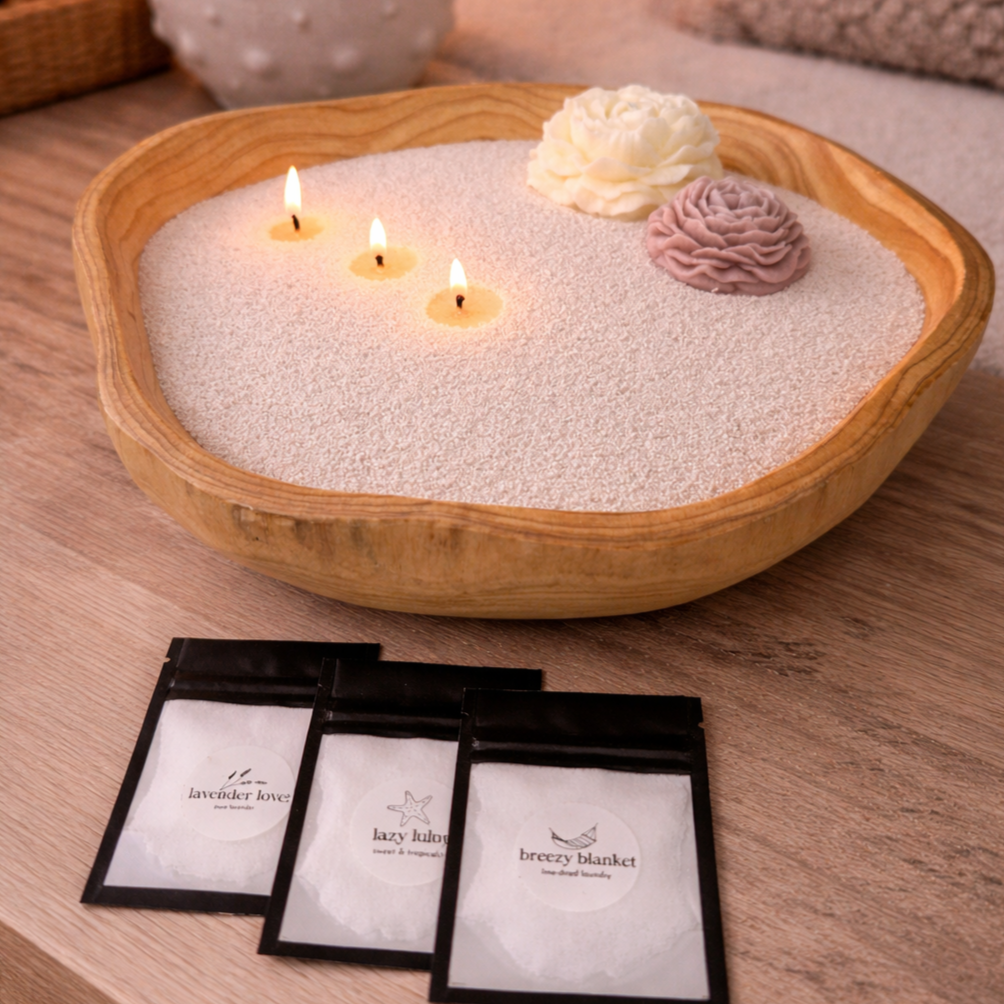 Lifestyle image featuring a handcrafted wood bowl candle filled with pearled wax, accented with decorative wax flowers and multiple lit wicks, accompanied by scent sample packets, styled in a warm, natural setting to highlight customization, fragrance variety, and a cozy, artisanal aesthetic.