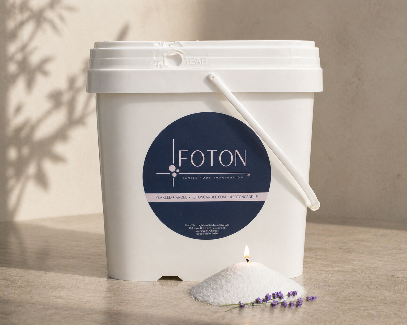 Large white Foton Pearled Candle bucket, shown on a neutral surface beside a small mound of white wax pearls with a lit wick and lavender sprig, in soft natural lighting.