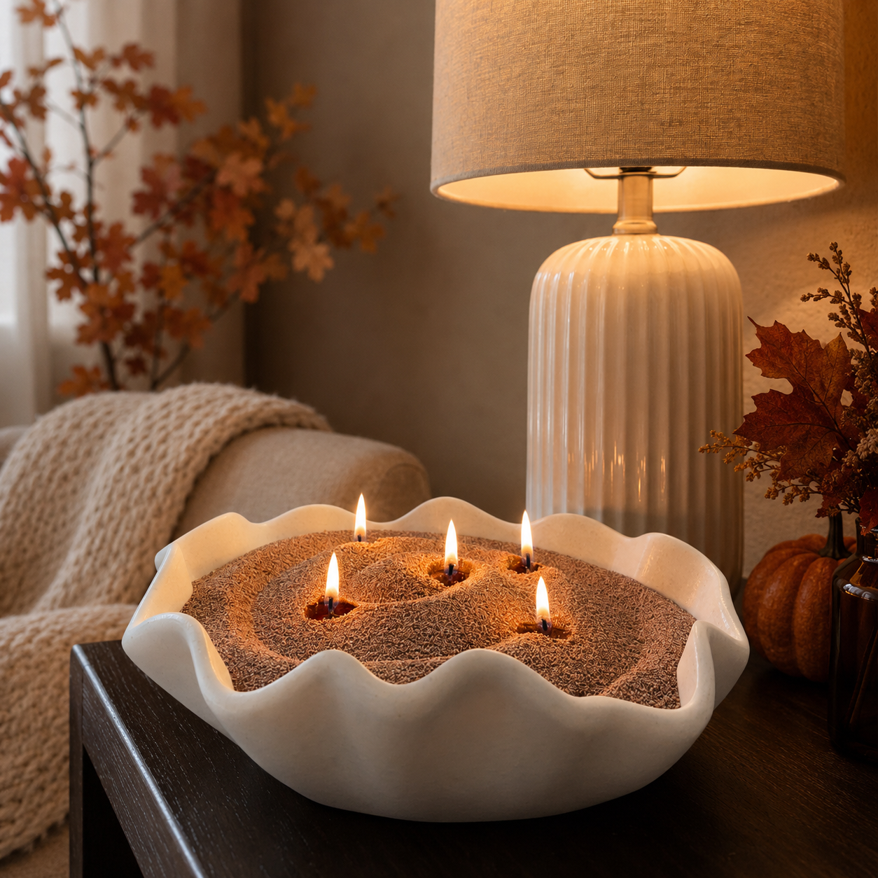 White scalloped ceramic bowl filled with Caramel Cashmere pearled candle wax and five glowing flames, styled on a dark wood side table beside a warm table lamp. Cozy fall accents, including pumpkins, autumn leaves, and soft knit textures, create an inviting seasonal atmosphere.