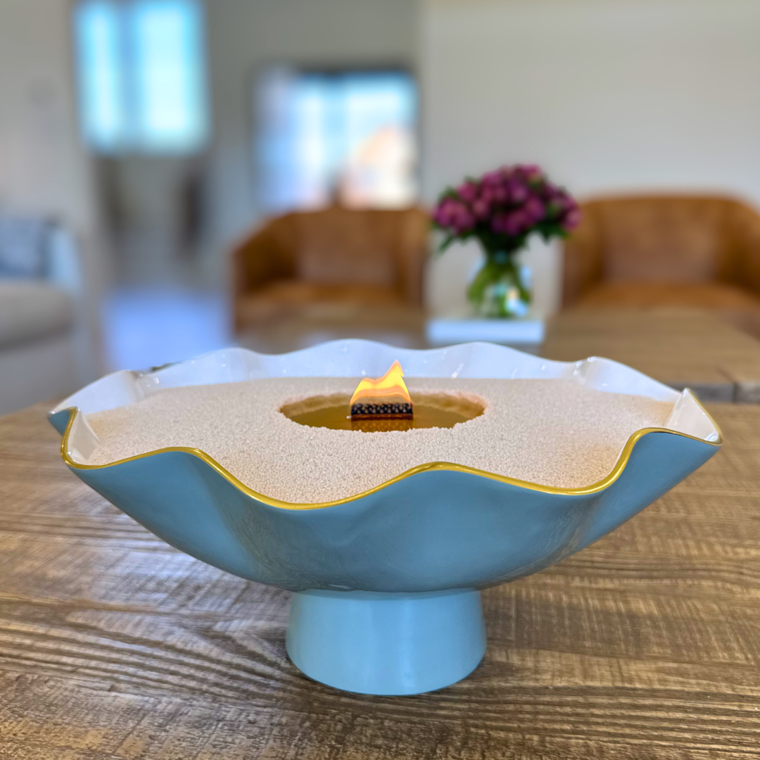 Decorative bowl candle made with candle pearls and a wooden wick, set on a table in a home setting with blurred seating and flowers in the background.