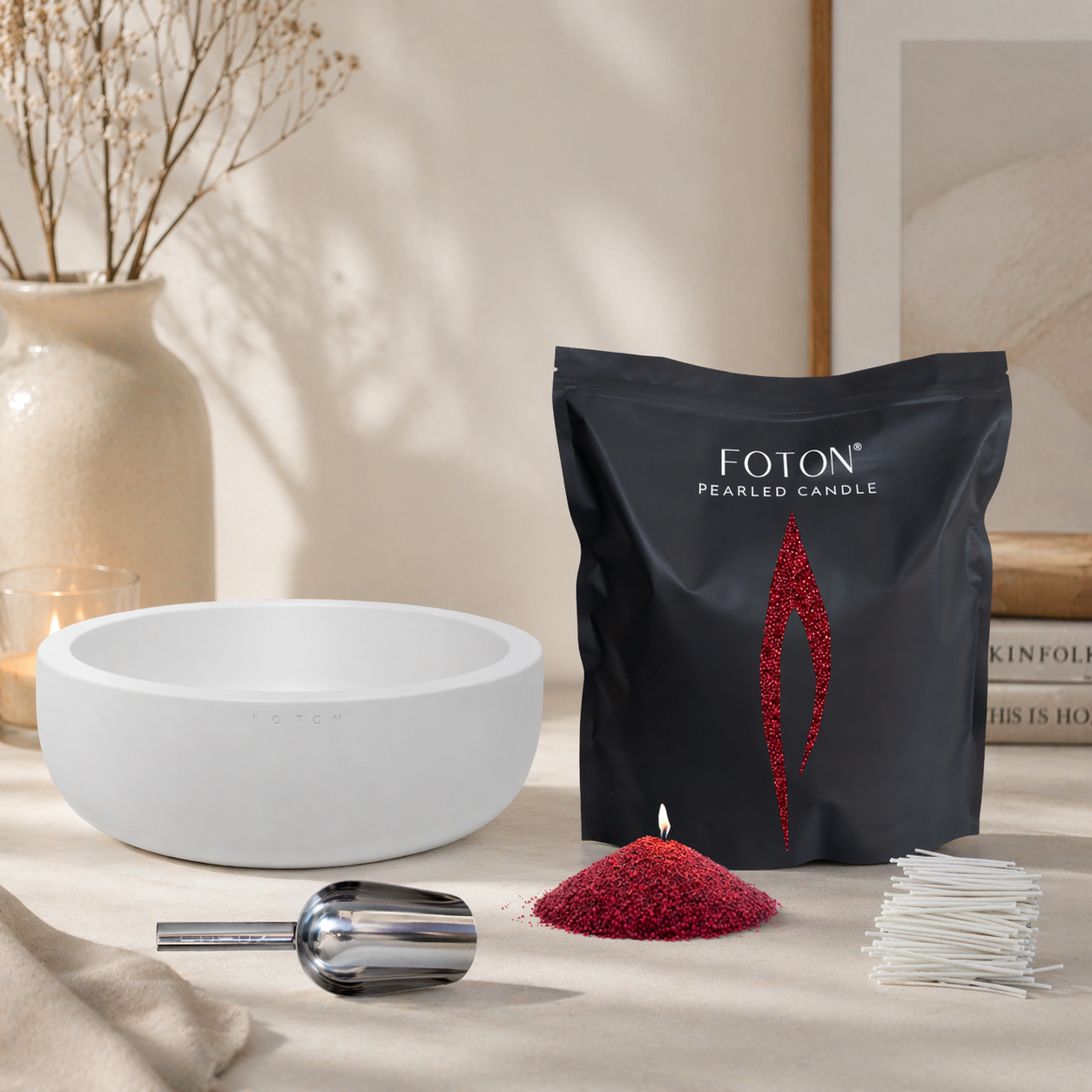 Minimalist lifestyle scene featuring a candle bundle arranged on a neutral tabletop. A Luxe white bowl sits to the left, a black Grande bag stands upright to the right, and in front of it is a small pile of maroon wax pearls with a lit wick. A metal scoop lies horizontally in the foreground, and a bundle of white wicks is stacked on the right. Soft natural light casts gentle shadows, with a ceramic vase holding dried branches and a lit candle in the background.
