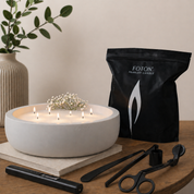 Cozy living room scene showing a large white Luxe concrete bowl candle with multiple lit wicks and baby breath flowers, set on a wooden table. A black Foton Pearled Candle Grande bag and candle care tools including a lighter, wick trimmer, dipper, and snuffer are arranged beside it, with warm ambient lighting and home decor in the background.