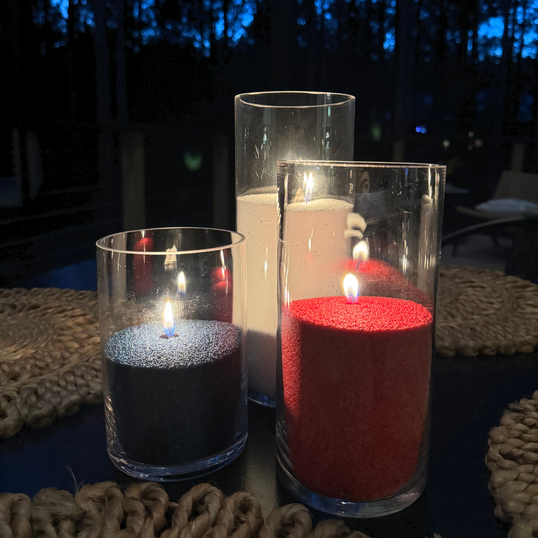 Patriotic-themed pearled candles displayed in tall clear glass vessels at night, featuring red, white, and blue wax with glowing flames on a dark outdoor table surrounded by woven placemats and a softly lit forest background.