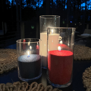 Patriotic-themed pearled candles displayed in tall clear glass vessels at night, featuring red, white, and blue wax with glowing flames on a dark outdoor table surrounded by woven placemats and a softly lit forest background.