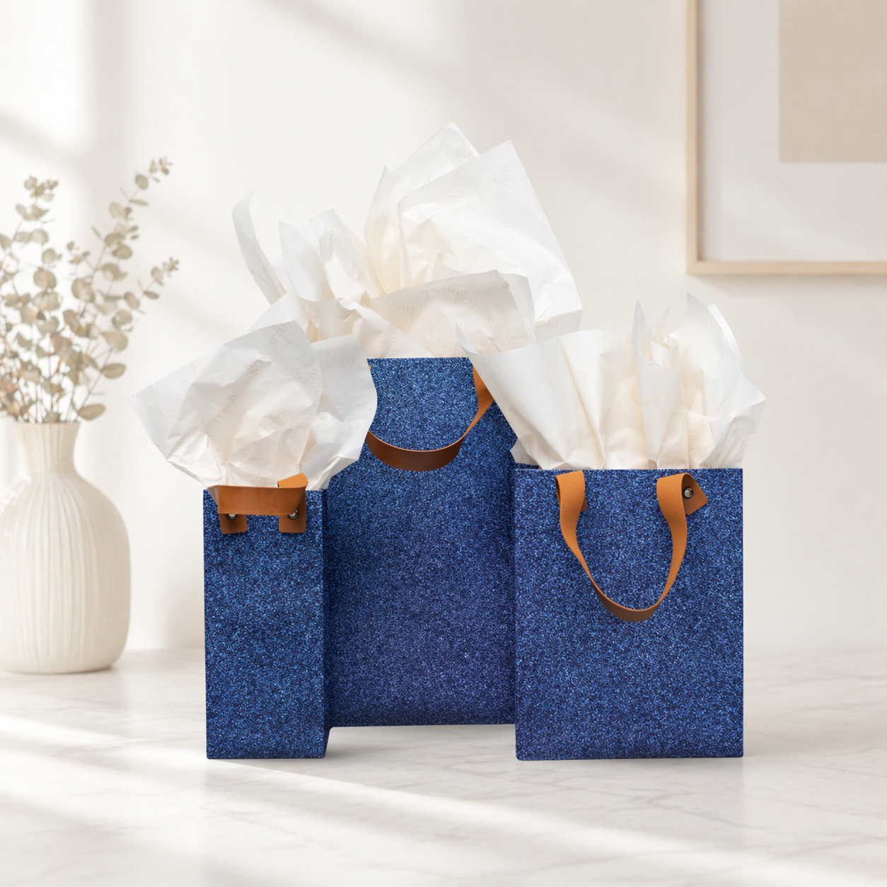 Three blue pearl-textured gift bags with tan leather handles, filled with light beige tissue paper, arranged on a light surface with a decorative vase and soft neutral background.