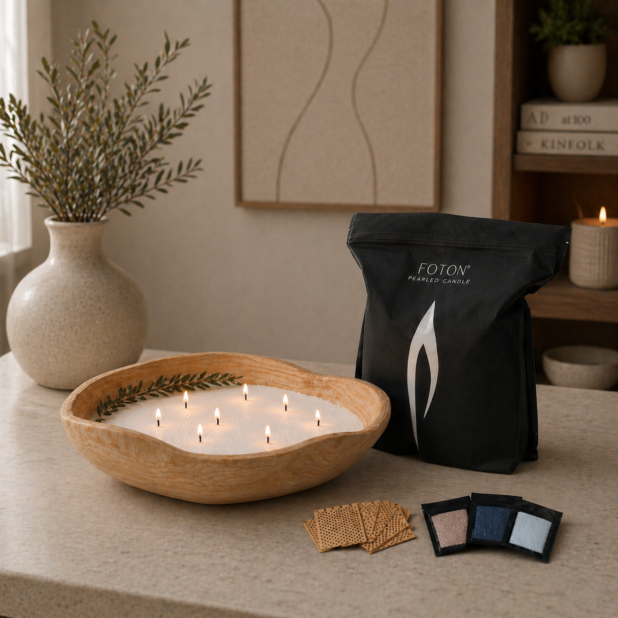 Styled home scene featuring a large lit candle with multiple small flames in a handcrafted wooden bowl on a countertop. Next to it is a black Foton Pearled Candle grande bag, along with XL wooden wicks and three sample packs. The background includes soft decor such as a ceramic vase with greenery, framed wall art, and warm ambient lighting.