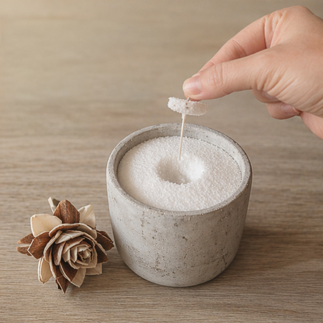 Hand placing a fresh wick into a small concrete candle filled with white wax pearls, creating a small indentation in the surface, with a decorative flower beside it.
