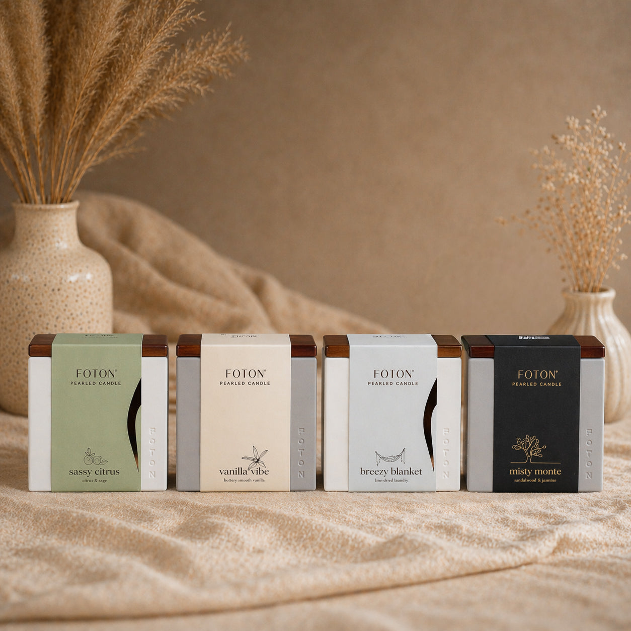 A clean, styled product image featuring four Foton Le Cube candles arranged side by side, each representing a different scent: Sassy Citrus, Vanilla Vibe, Breezy Blanket, and Misty Monte. The neutral, textured background with soft textiles and dried florals creates a warm, elevated aesthetic that highlights the variety and sophistication of the collection.