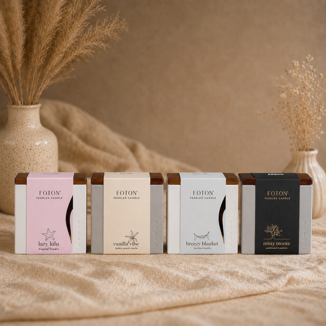 A clean, styled product image featuring four Foton Le Cube candles arranged side by side, each representing a different scent: Lazy Lulu, Vanilla Vibe, Breezy Blanket, and Misty Monte. The neutral, textured background with soft textiles and dried florals creates a warm, elevated aesthetic that highlights the variety and sophistication of the collection.