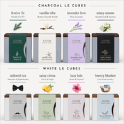 Grid display of Le Cube candle scent options, featuring two colors: Charcoal and White. The Charcoal line includes festive fir (freshly cut fir), vanilla vibe (buttery smooth vanilla), lavender love (pure lavender), and misty monte (sandalwood & jasmine). The White line includes tailored tux (masculine & sophisticated), sassy citrus (citrus & sage), lazy lulu (sweet & tropical), and breezy blanket (line-dried laundry), each shown with corresponding packaging.