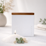 Lifestyle product image of the Le Cube candle in a clean white vessel, styled with soft greenery and neutral tones to evoke a calm, minimalist, spa-like ambiance