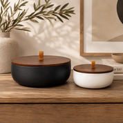 Matte black and white Luxe concrete candle bowls with matching wooden lids displayed on a warm wood console table in a modern home. The bowls are styled with neutral décor, ceramic vases, framed artwork, and soft natural light to highlight their timeless, minimalist design.
