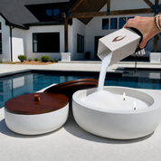White Luxe Concrete Bowl being refilled with Foton Candle pearls beside a swimming pool. The image demonstrates the easy refill process and the reusable design of the concrete candle vessel.