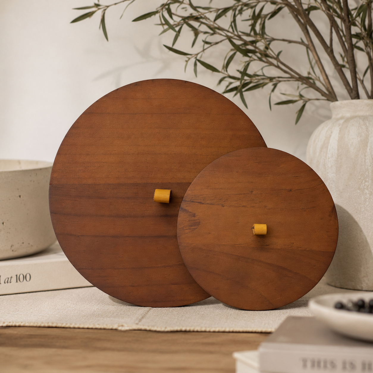 Large and small round wooden candle lids displayed upright on a neutral tabletop, showcasing their rich natural wood grain and minimalist design. The lids are styled with ceramic décor, books, and greenery in a warm, modern interior.