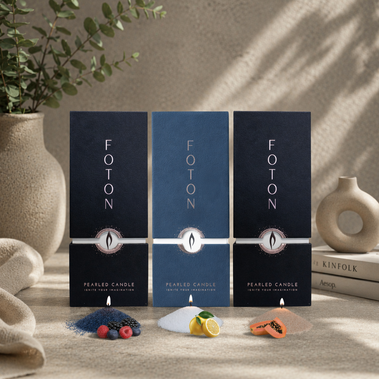 Three Foton pearled candle boxes displayed side by side in a neutral, styled setting. In front of each box are small mounds of wax with lit wicks and scent cues: berries for a fruity scent, lemon for a citrus scent, and papaya for a tropical scent. Decorative elements like vases, greenery, and books create a soft, modern background.