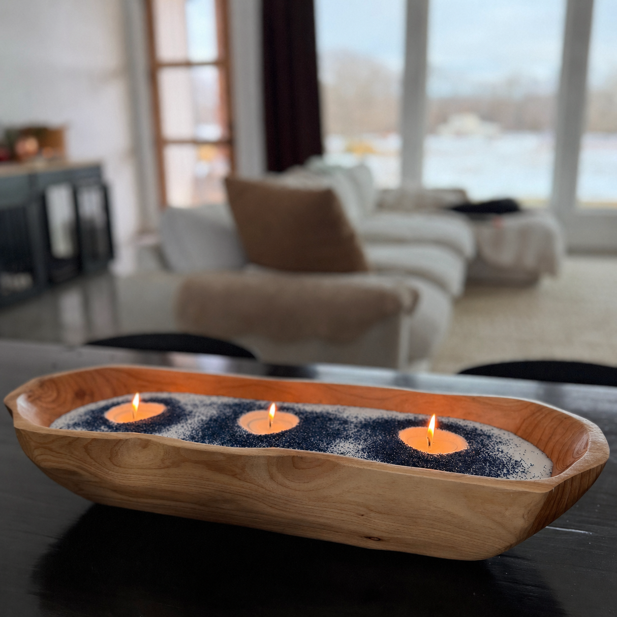 Handcrafted wood bowl in a modern dough bowl shape candle with three lit flames arranged in a row, featuring navy blue decorative wax accents around each wick. The candle sits on a dark table in a softly blurred living room with a couch, and large windows in the background.