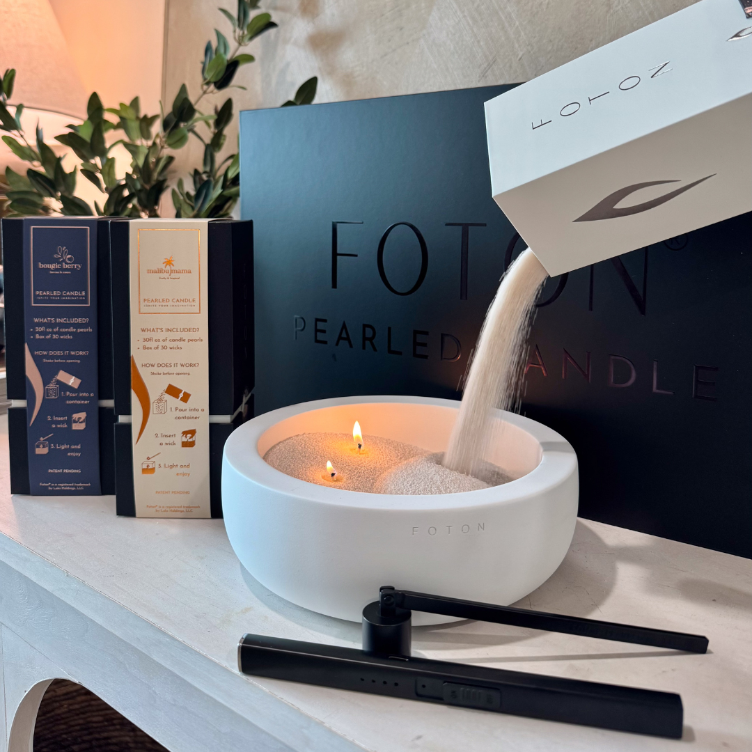 Close-up of white candle pearls being poured from a box into a white Luxe concrete bowl candle with lit wicks, with two 18oz limited edition candles, a black electric lighter, snuffer, and a Foton luxury gift box in the background.