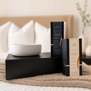 White Luxe concrete candle bowl resting on a closed black Foton luxury gift box on a bed, next to two 18oz limited edition candle boxes labeled Bougie Berry and Malibu Mama, with a soft neutral bedroom background.