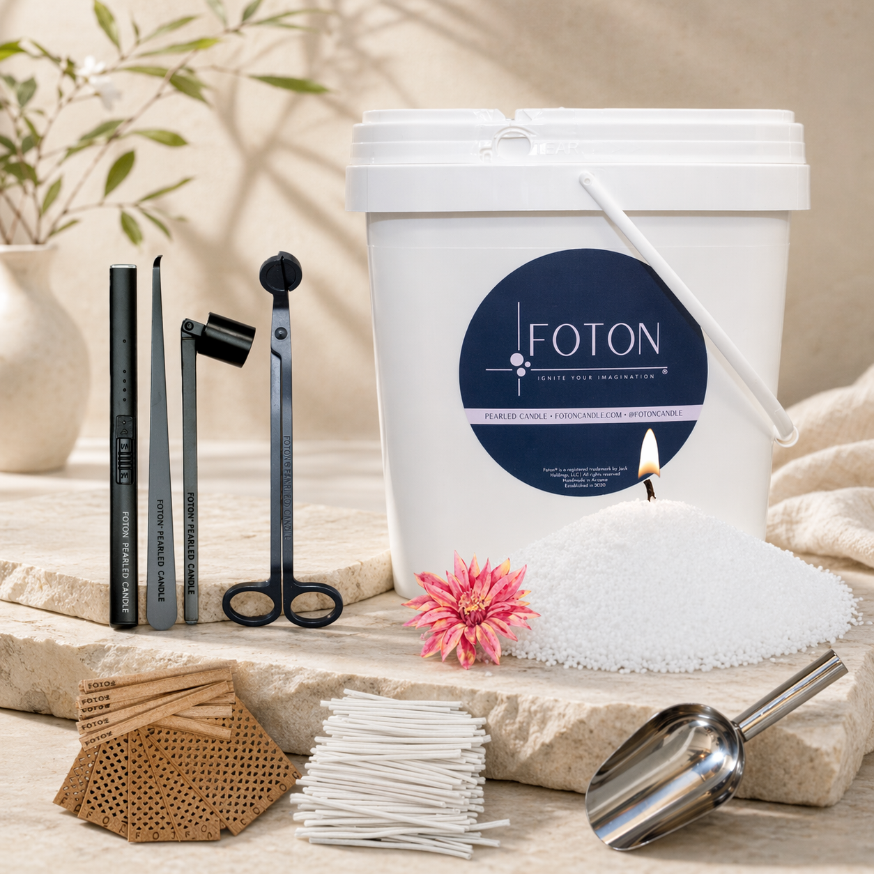 Styled product display of a Foton candl XL-kit on a neutral stone surface. A white bucket with a “FOTON” label is shown alongside a mound of white pearled wax with a single lit wick. Arranged around it are candle care tools including an electric lighter, wick dipper, wick trimmer, snuffer, a metal scoop, bundles of cotton and wooden wicks, accented by a pink flower and soft, natural decor in the background.