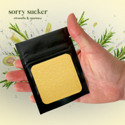 Promotional product image for the “Sorry Sucker” citronella and rosemary scent sample featuring a hand holding a black pouch filled with pale yellow pearled candle wax against a fresh botanical background with rosemary sprigs and citronella imagery.