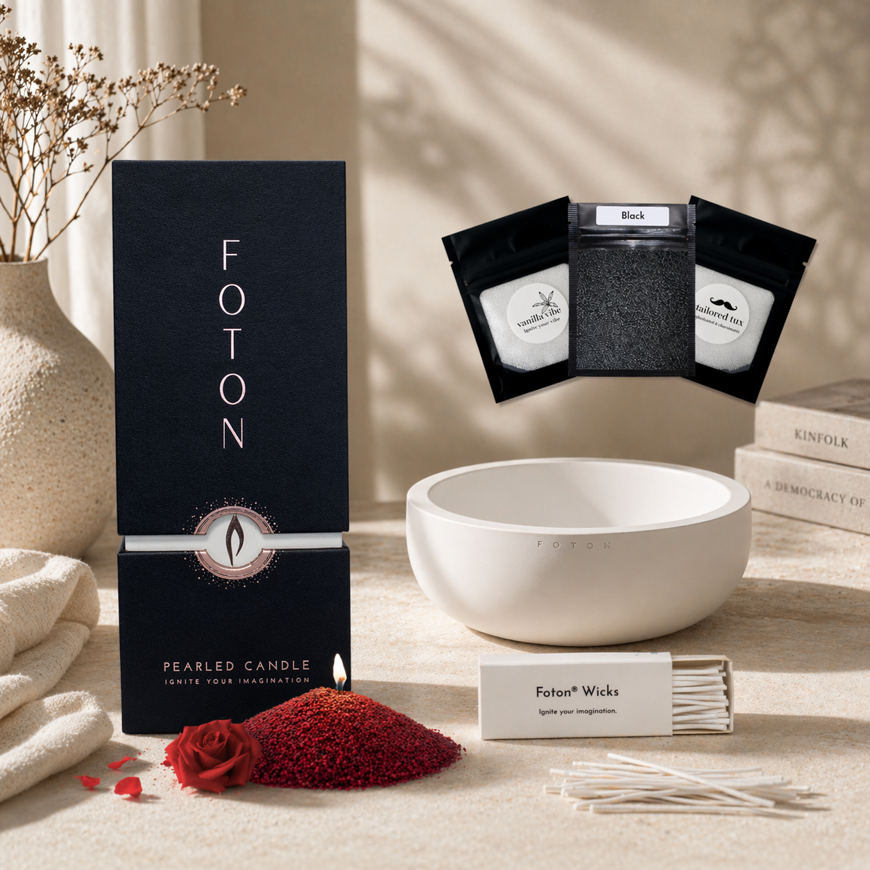 A Foton pearled candle 18oz box arranged on a light beige table in warm, natural lighting, featuring a white Luxe candle vessel, a box of wicks with loose wicks placed on the table, and three small black sample pouches above. In front, a small pile of maroon candle pearls with a lit wick sits next to a red rose, while soft decor elements like a vase, books, and fabric create a calm, spa-like background.