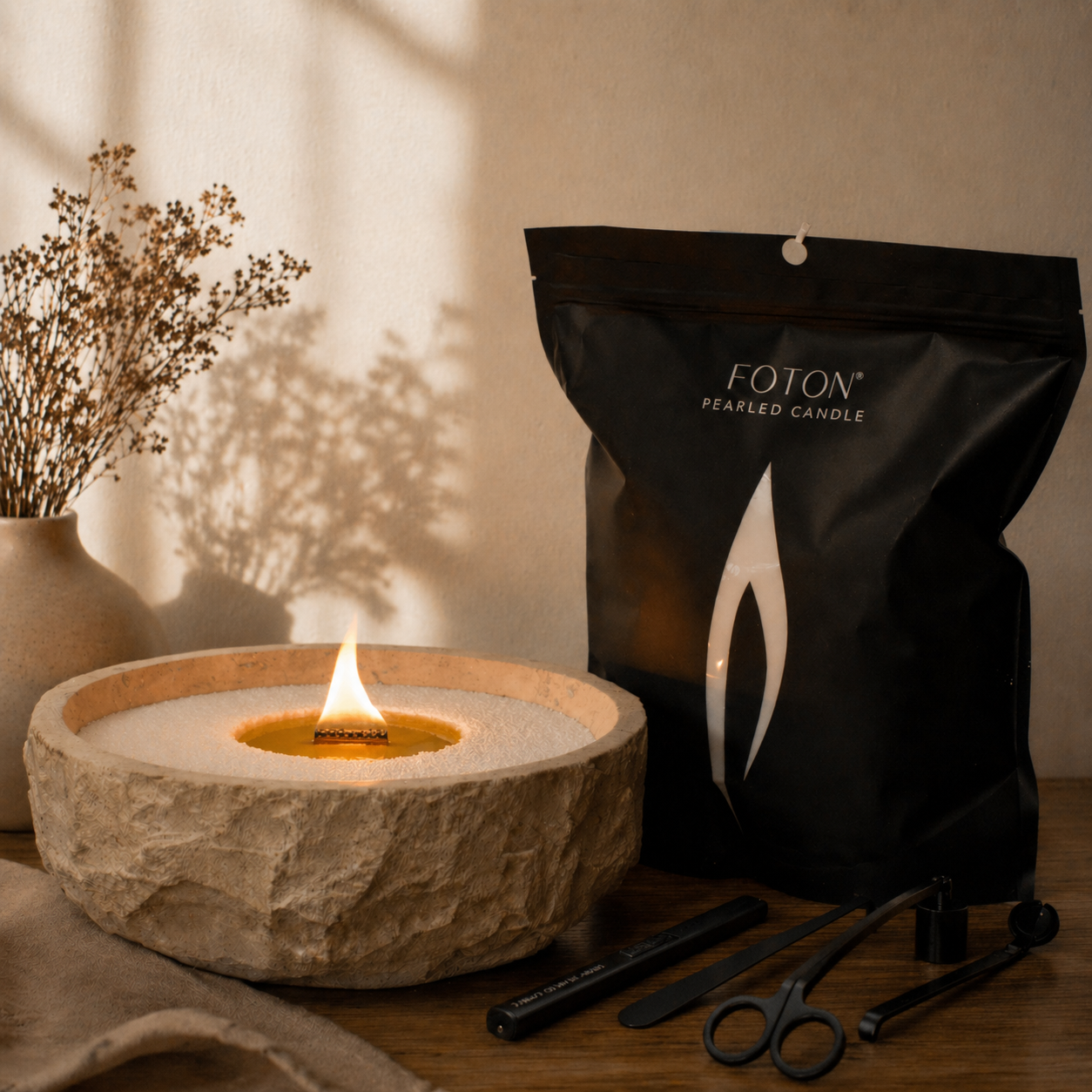Styled product scene showing a lit candle in a textured stone bowl with a wooden wick flame, placed on a wooden surface. Beside it is a black Foton Pearled Candle grande bag and a set of candle care tools including an electric lighter, wick trimmer, wick dipper, and flame snuffer, with soft natural lighting and dried floral decor in the background.