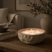 A warm, inviting lifestyle shot featuring a multi-wick Foton candle set in a textured stone vessel. The soft glow of four flames creates a cozy ambiance, complementing the earthy tones of the wooden surface, neutral textiles, and natural decor elements. Styled to evoke comfort and calm, the scene highlights the candle as a statement piece for relaxed, elevated home living.