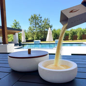 Outdoor lifestyle scene featuring a white Luxe concrete candle vessel being filled with pale yellow pearled candle wax from branded packaging beside a matching lidded vessel, set on a patio table overlooking a swimming pool and sunny backyard.