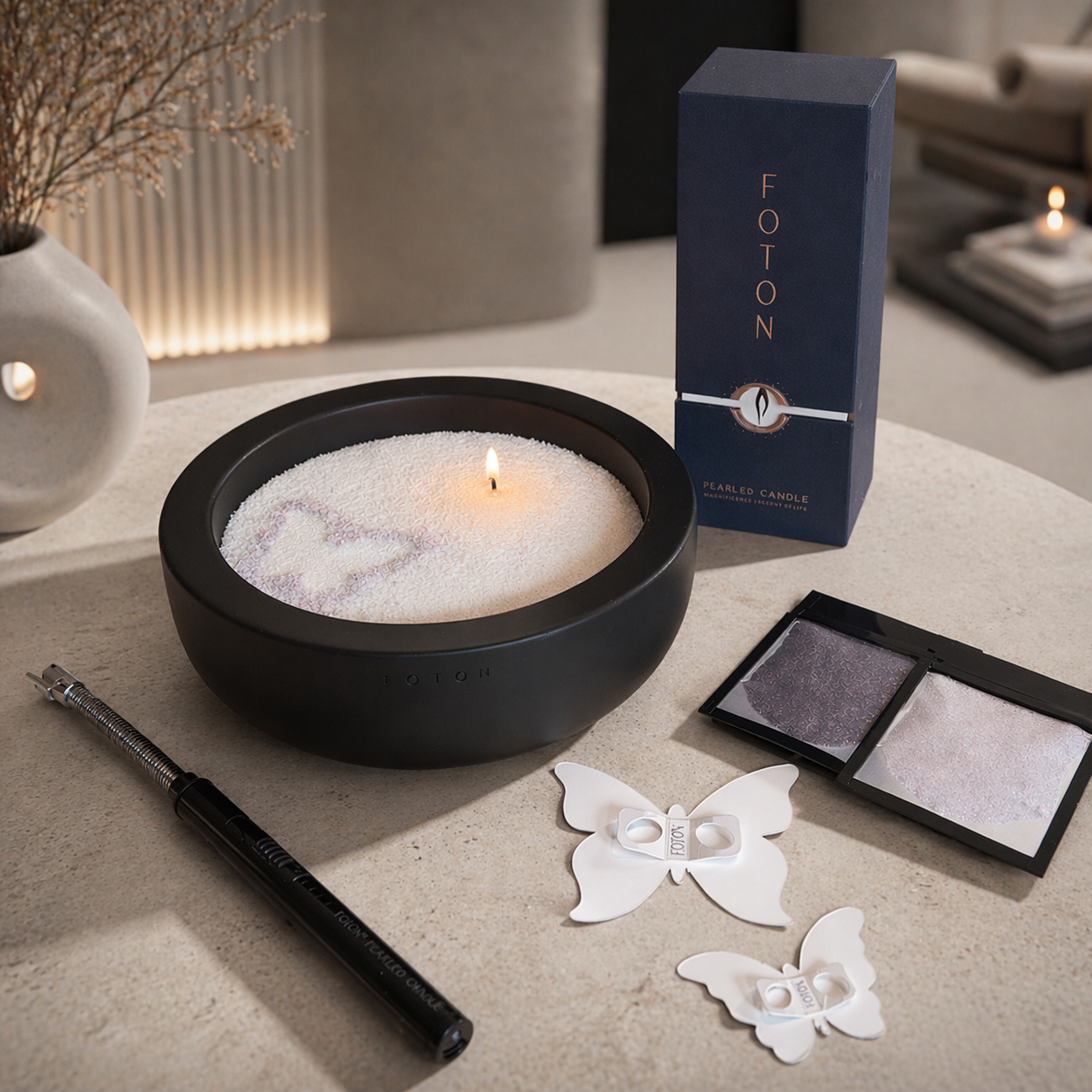 Foton candle set displayed on a stone table, featuring a black Luxe concrete vessel filled with white pearled wax and a single lit wick with a faint butterfly design. Accompanying items include a navy 18oz Foton box, two packets of colored wax samples in amethyst and lilac, two butterfly-shaped stencils, and a black electric lighter placed beside the candle.