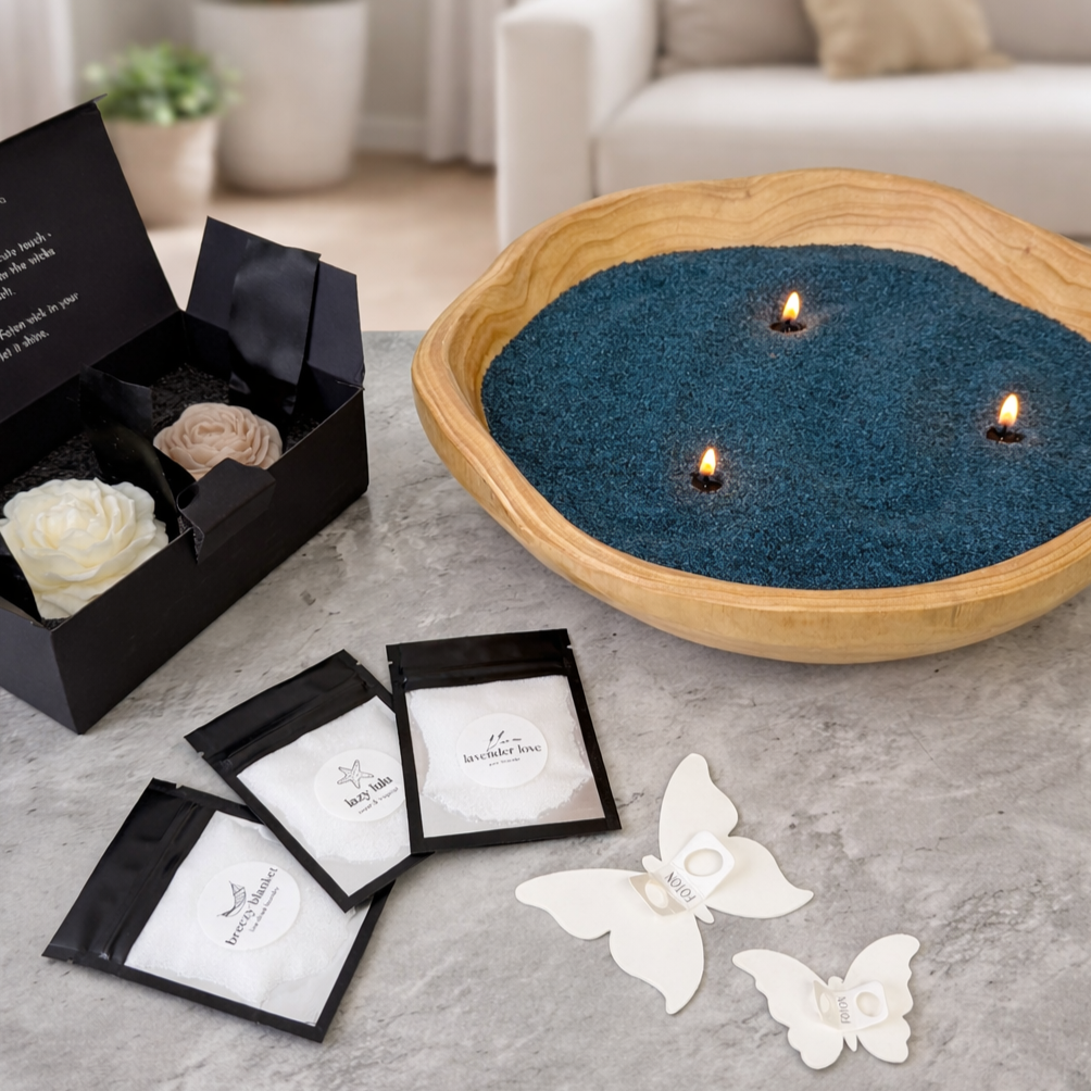 Lifestyle product image of a Foton pearled candle in a handcrafted wooden bowl, filled with deep blue pearled wax and lit with multiple wicks. The scene includes an open gift box with wax flower accents, scent sample packets, and butterfly stencils, highlighting the complete customizable candle kit in a clean, modern home setting.