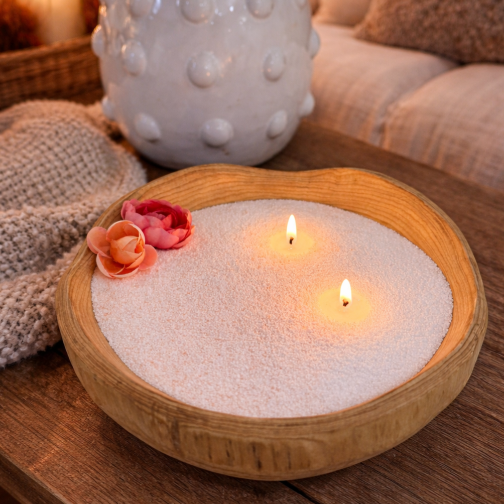 A cozy, warm-toned lifestyle shot of a pearled candle set in a handcrafted wood bowl with softly rounded edges. The candle features two gently glowing wicks surrounded by smooth, sand-like wax, accented with delicate pink and coral flowers for a soft, feminine touch. Set on a wooden table with a knitted throw and neutral décor in the background, the scene evokes a calm, inviting, and homey ambiance perfect for relaxation or gifting.
