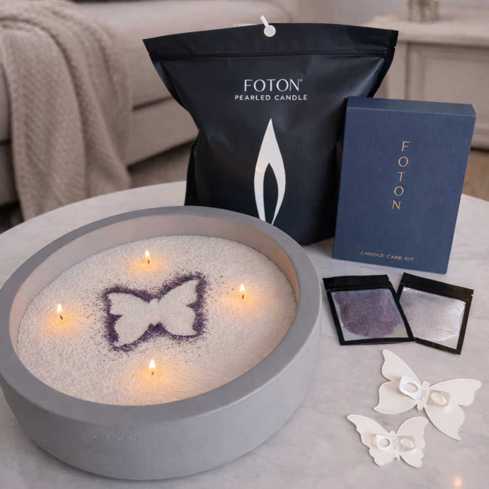 Foton candle set displayed on a tabletop, featuring a round Luxe concrete vessel filled with white pearled wax and four lit wicks, decorated with a purple butterfly stencil design. Behind it are a grande bag of wax, and a navy candle care kit box. In front are butterfly-shaped stencils and small packets of colored wax for customization.