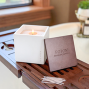 A cozy lifestyle image featuring a lit Foton Le Cube candle, paired with its matching lid and cotton wicks. Set on a wooden bath tray with soft natural light, the scene evokes a calm, spa-like atmosphere that highlights both the product’s elegance and everyday usability.
