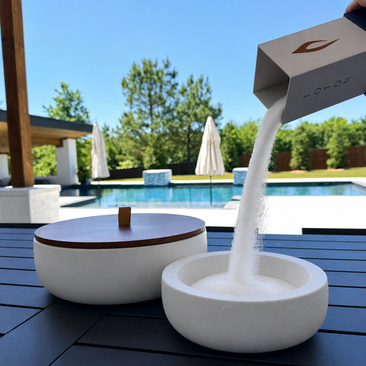 Outdoor lifestyle scene featuring a white Luxe concrete candle vessel being filled with white pearled candle wax from branded packaging beside a matching lidded vessel, set on a patio table overlooking a swimming pool and sunny backyard.