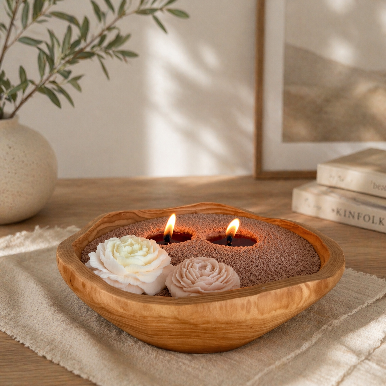 Pearled candle in a handcrafted wood bowl with two lit flames set in desert rose colored wax pearls, accented with two flower-shaped wax pieces, placed on a soft cloth with books and a vase in a warm, softly lit home setting.