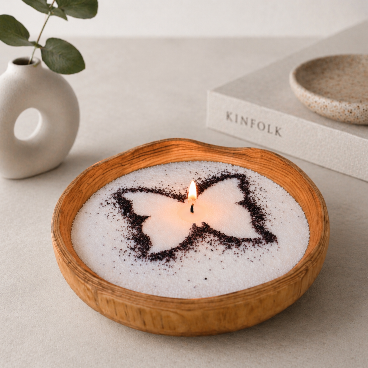 Petite handcrafted wood bowl candle filled with white pearled wax and a single lit wick, featuring a dark butterfly stencil design on the surface. The candle sits on a neutral tabletop beside a small ceramic vase with greenery and a book, creating a soft, minimalist home setting.