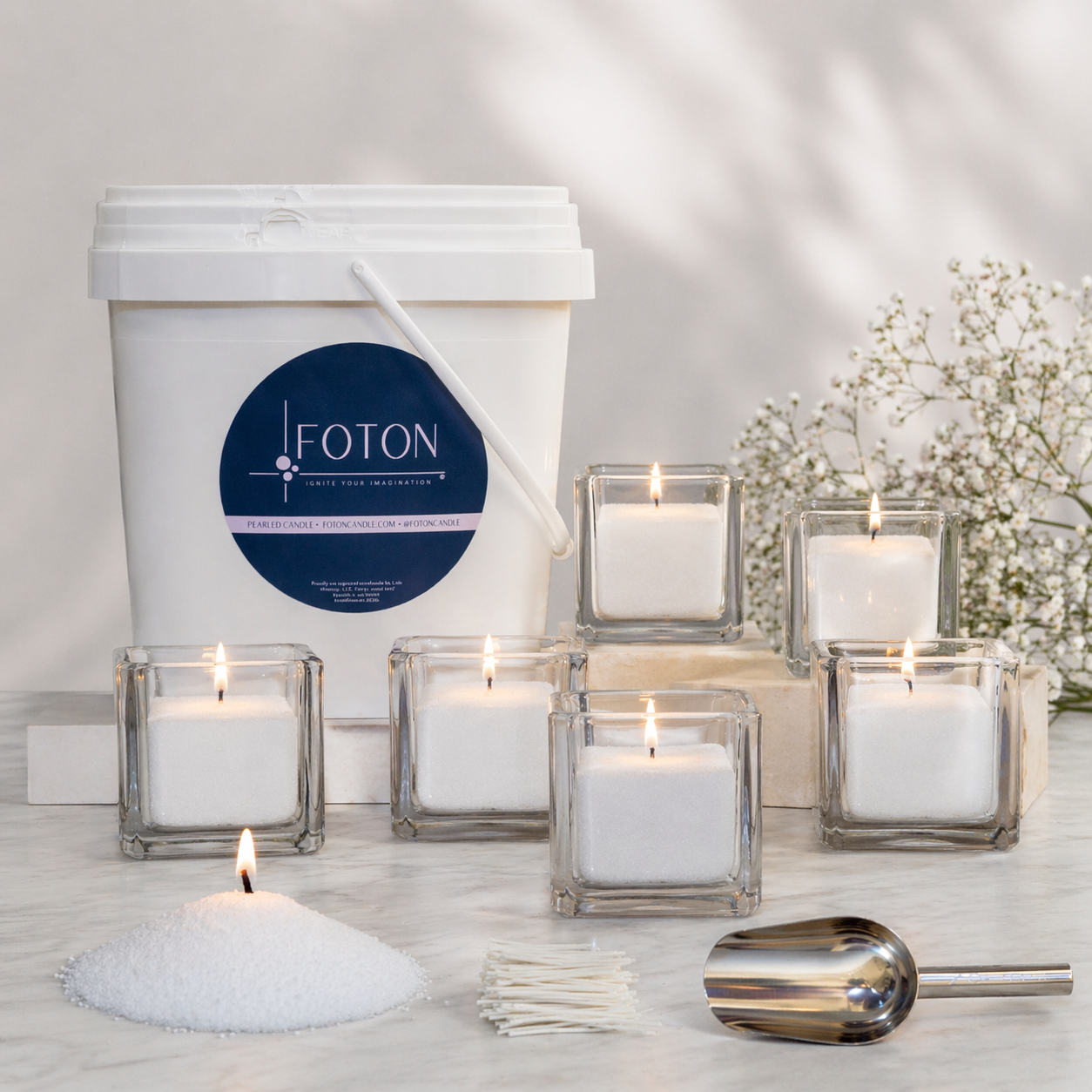 FOTON pearled candle kit display featuring a large white bucket of wax, 6 square glass candle holders with lit candles, loose pearled wax, wicks, and a metal scoop, arranged on a light surface with soft floral accents in the background.